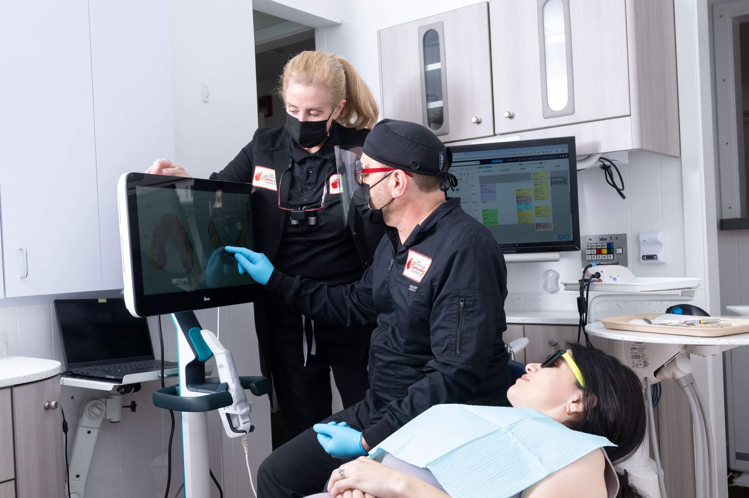 Two dental professionals in masks and scrubs show a patient her 3D dental scan on a monitor in a modern treatment room.
