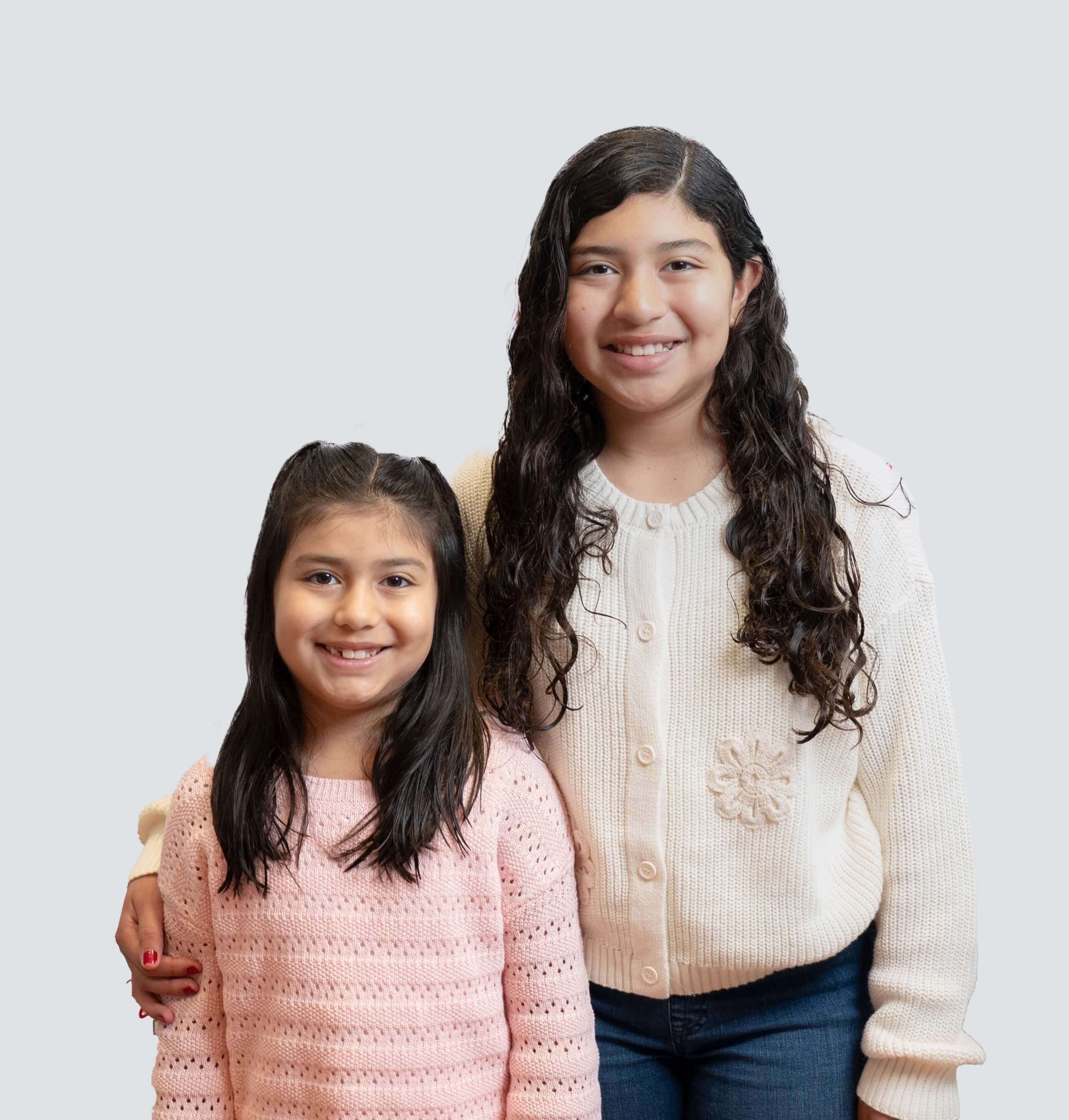 Two smiling girls, an older sister with long dark curly hair and a younger sister with dark hair in pigtails, representing our family-friendly dental care.